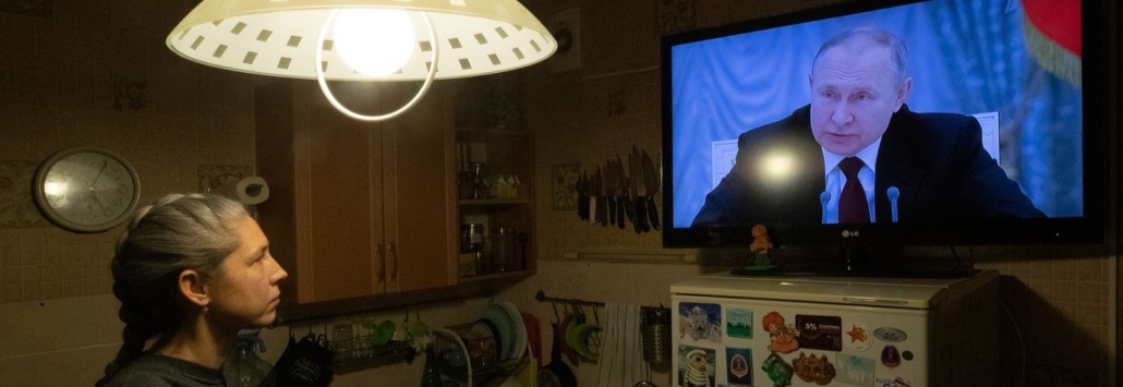 Photo d'un homme assis dans son salon, dans l'obscurité, en train de regarder Vladimir Poutine à la télévision